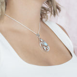 The picture shows a model wearing a 925 sterling silver teardrop shaped pendant with 3 sea turtles in the center with cubic zirconia and larimar.