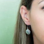 In this picture there is a close-up of a model with brown hair wearing sterling silver oyster shell dangle earrings.
