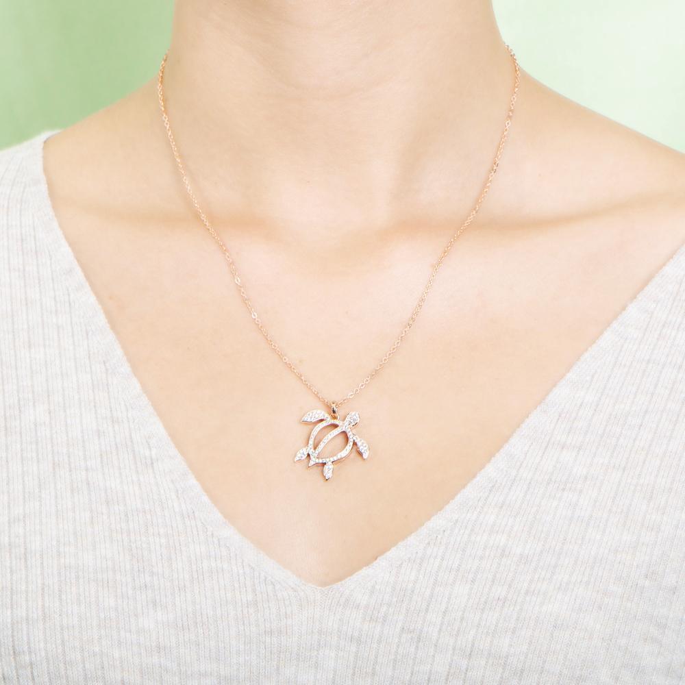 In this photo there is a model with blonde hair and a white shirt, wearing a rose gold sea turtle pendant with topaz gemstones.