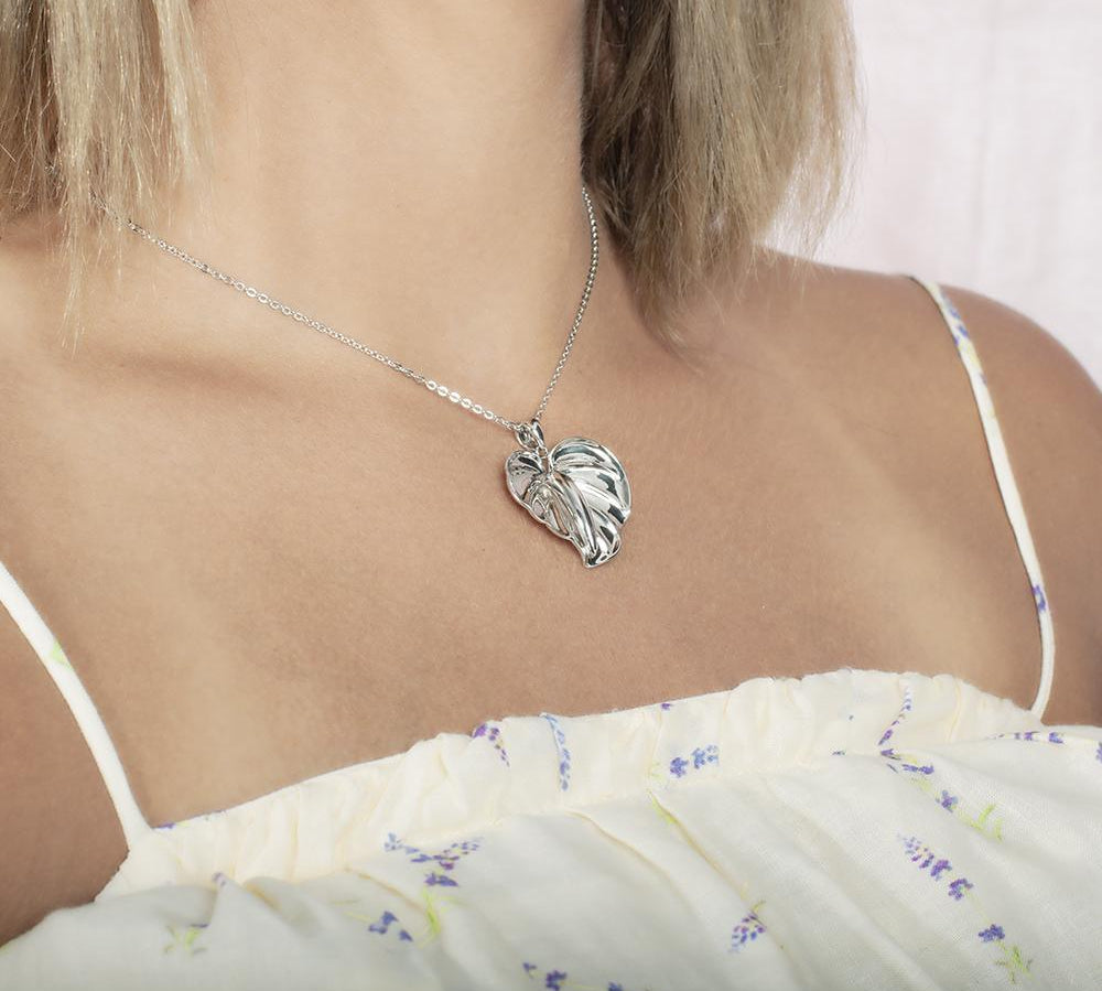 In this photo there is a model turned to the right with blonde hair and a white shirt with purple flowers, wearing a sterling silver anthurium pendant.