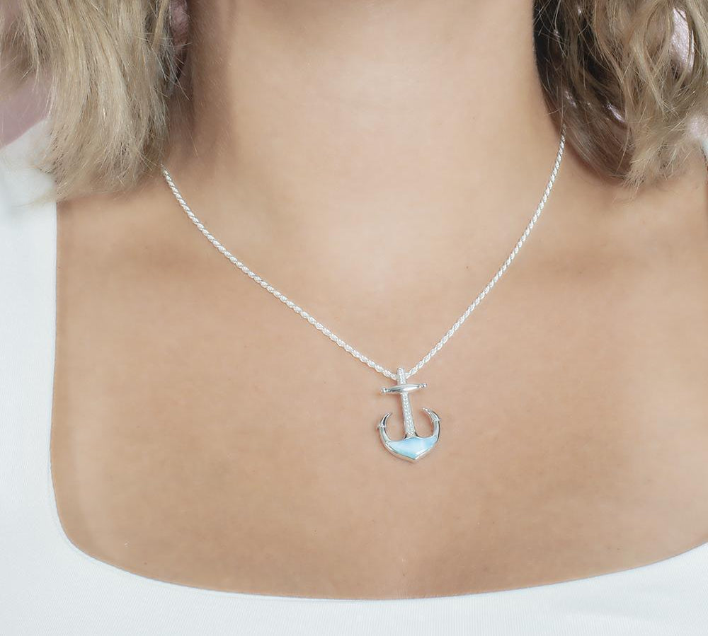 In this photo there is a model with blonde hair and a white shirt, wearing a sterling silver anchor pendant with blue larimar and topaz gemstones.