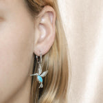 In this photo there is a model wearing 925 sterling silver flying hummingbird lever-back earrings with blue larimar and topaz gemstones.