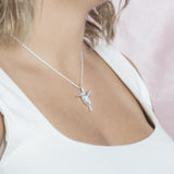In this photo there is a model with blonde hair and a white shirt turned to the right, wearing a sterling silver flying hummingbird pendant with topaz and one blue larimar gemstone.