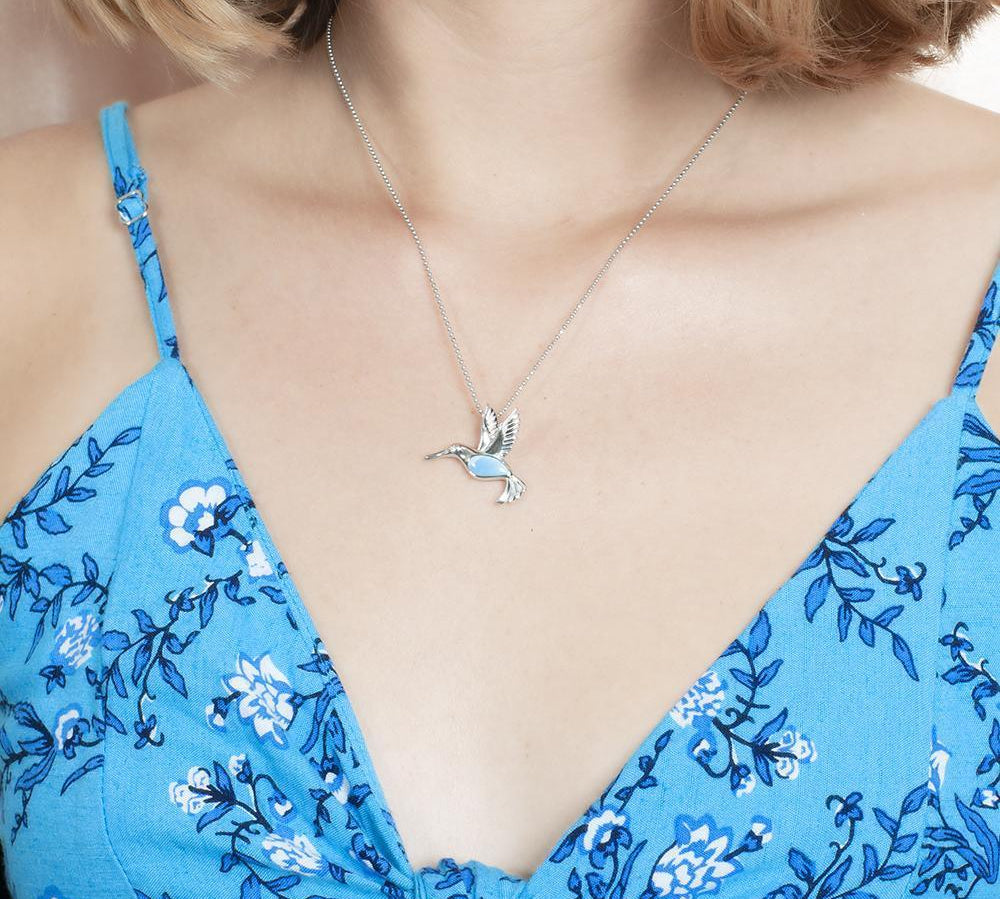 In this photo there is a model with blonde hair and a blue dress with flowers, wearing a sterling silver hummingbird pendant with one blue larimar gemstone.