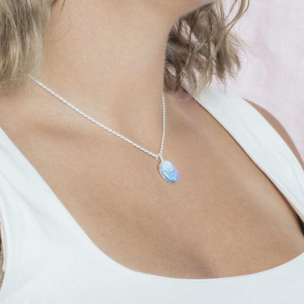 In this photo there is a model turned to the right with blonde hair and a white shirt, wearing a sterling silver circle pendant with blue larimar, opalite, and topaz gemstones.