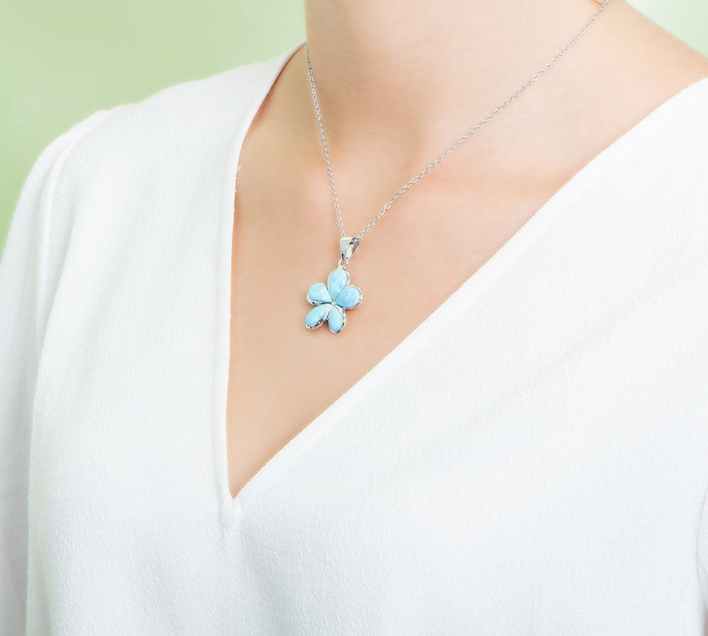In this photo there is a model with a white shirt turned to the left, wearing a medium sized sterling silver plumeria pendant with blue larimar gemstones.