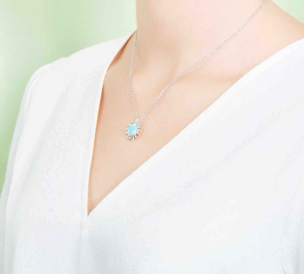 In this photo there is a model turned to the left with a white shirt, wearing a sterling silver sunflower pendant with one blue larimar gemstone.