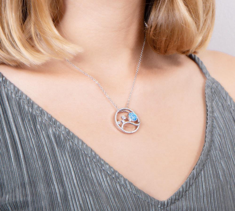 In this photo there is a model turned to the right with blonde hair and a gray shirt wearing a sterling silver circle pendant with blue opalite, aquamarine and cubic zirconia.