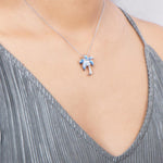 In this photo there is a model with a gray shirt turned slightly to the right, wearing a sterling silver palm tree pendant with blue opalite and three topaz gemstones.