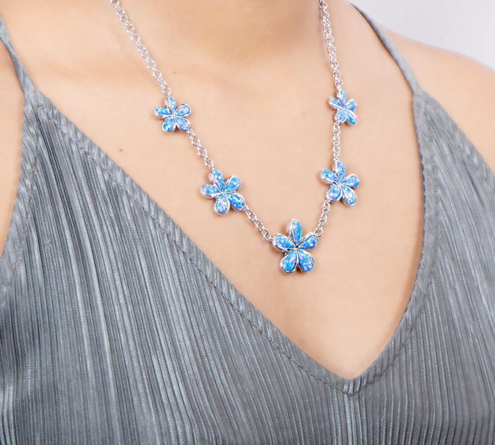 In this photo there is a model with a gray shirt wearing a sterling silver charm necklace with five plumeria flowers and blue opalite gemstones.