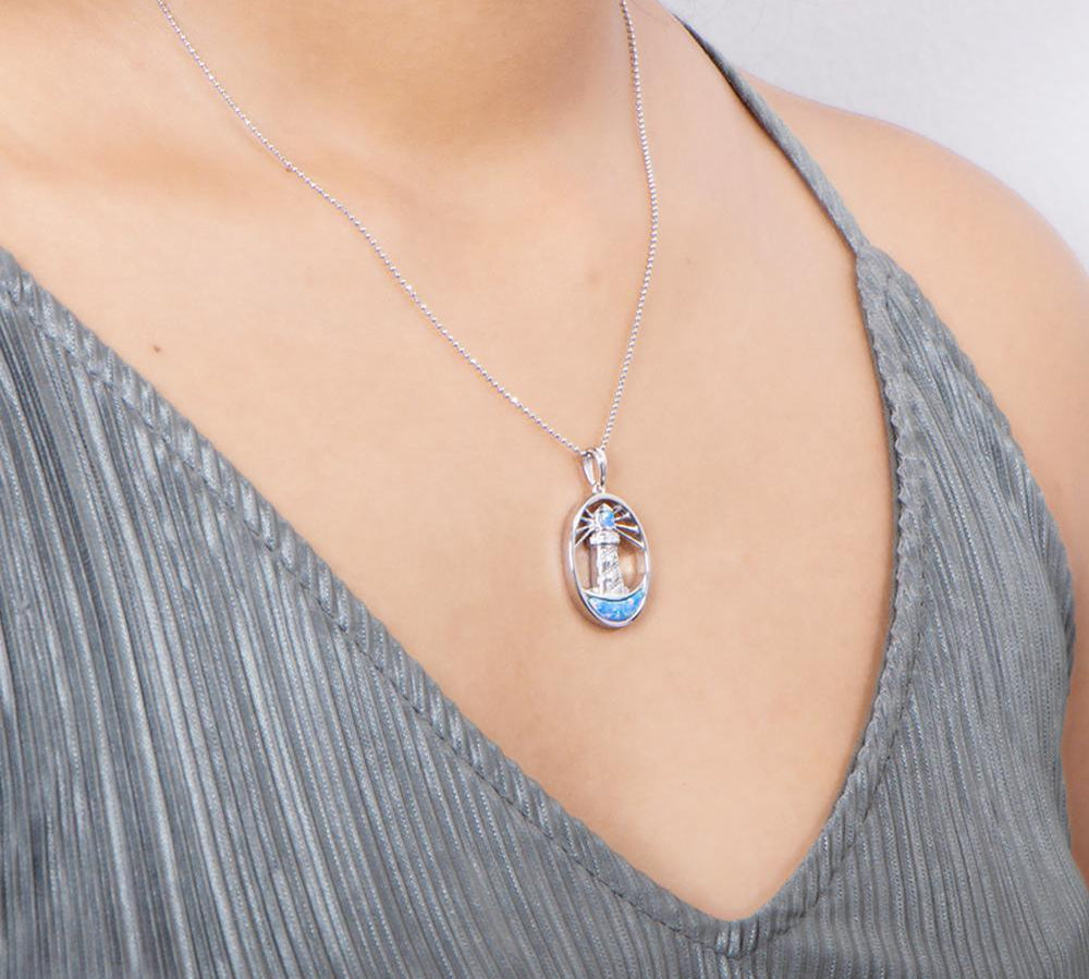 In this photo there is a model with a gray shirt turned slightly to the right, wearing a sterling silver shining lighthouse pendant with blue opalite and topaz gemstones.