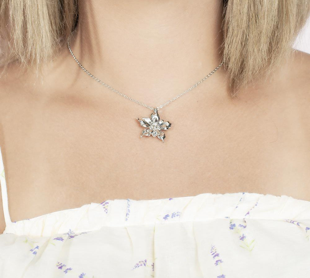 In this photo there is a model with blonde hair and white shirt with purple flowers, wearing a sterling silver orchid pendant.