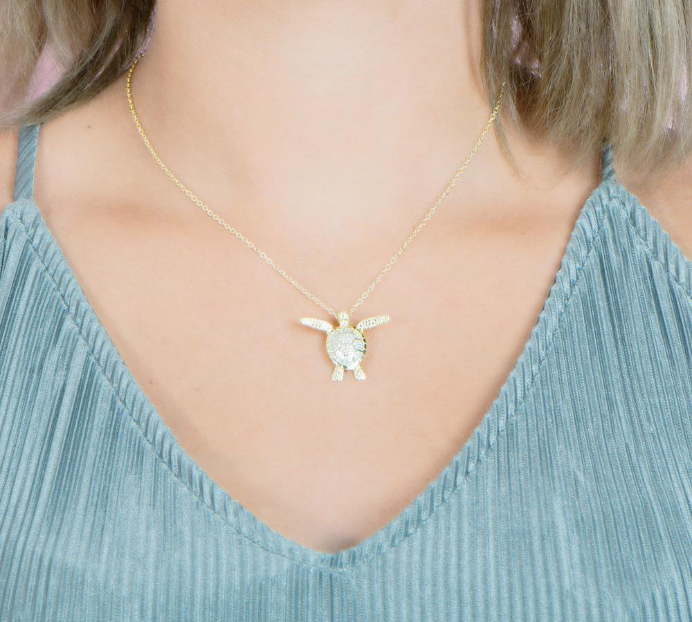 In this photo there is a model with blonde hair and a teal shirt, wearing a yellow gold sea turtle pendant with cubic zirconia.