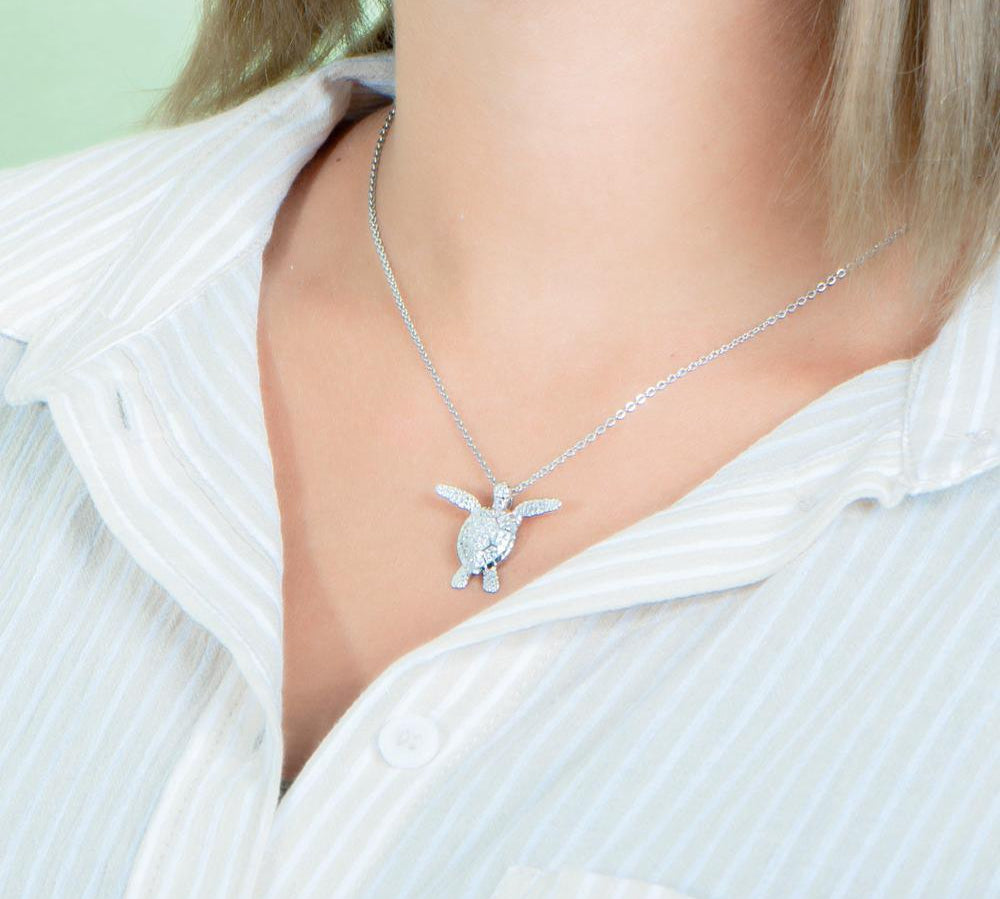 In this photo there is a model turned to the left with blonde hair and a tan and white striped shirt, wearing a white gold sea turtle pendant with cubic zirconia.