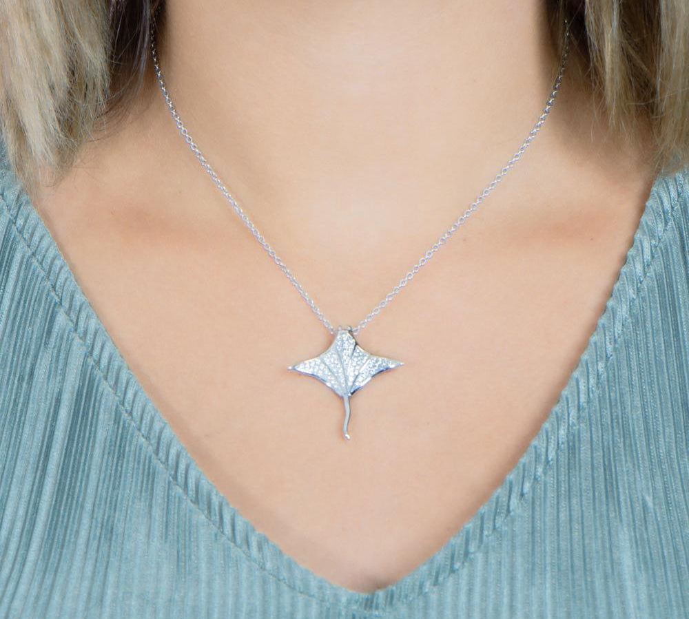 The picture shows a model wearing a 925 sterling silver, white gold vermeil, pavé manta ray pendant with topaz.