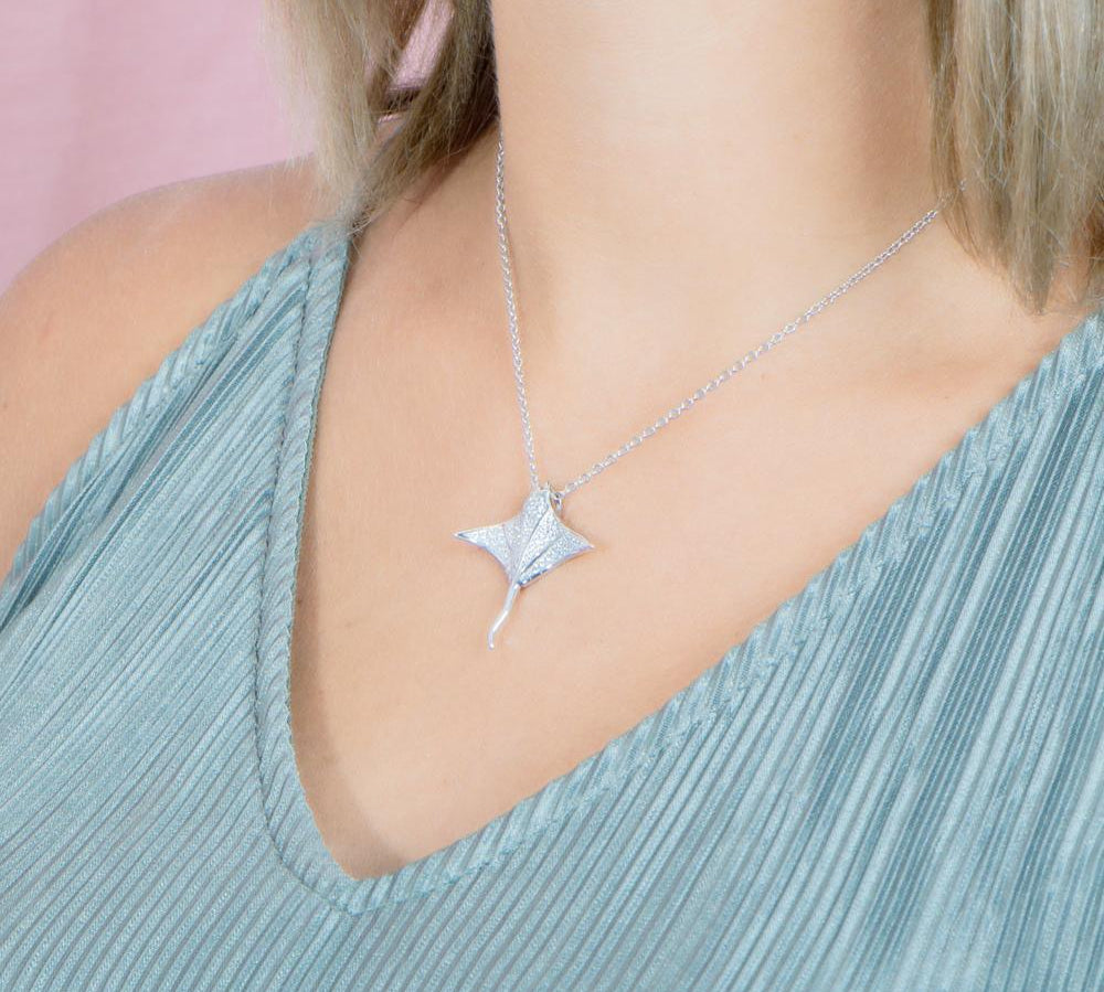 The picture shows a model wearing a 925 sterling silver, white gold vermeil, pavé manta ray pendant with topaz.