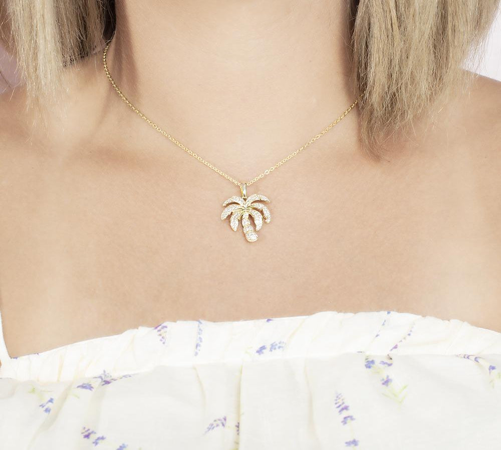 In this photo there is a model with blonde hair and a white shirt with flowers, wearing a yellow gold palm tree pendant with topaz gemstones.