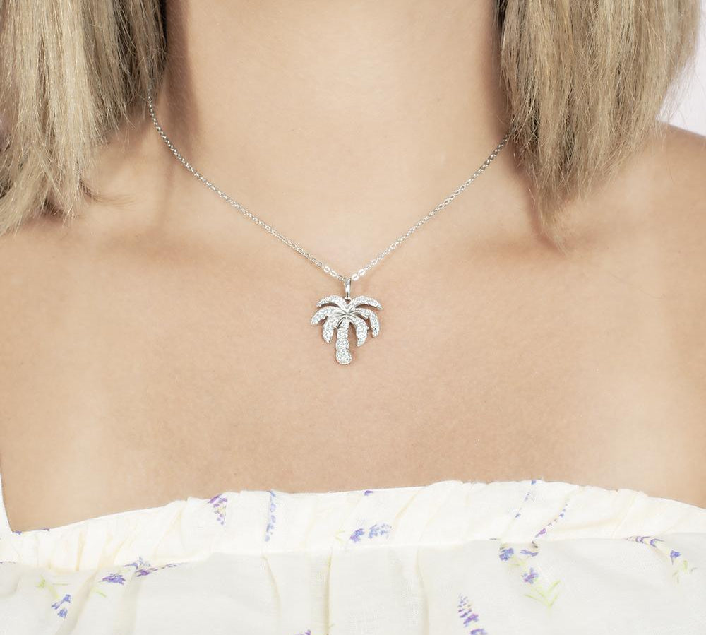 In this photo there is a model with blonde hair and a white shirt with flowers, wearing a white gold palm tree pendant with topaz gemstones.