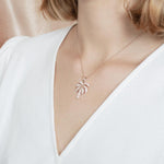 In this photo there is a model turned slightly to the left with blonde hair and a white shirt, wearing a rose gold palm tree pendant with topaz gemstones.