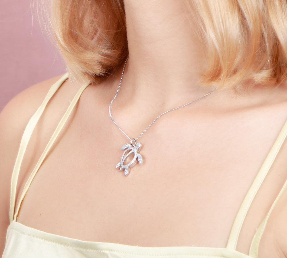 In this photo there is a model with blonde hair and a yellow shirt turned to the left, wearing a white gold sea turtle pendant with topaz gemstones.
