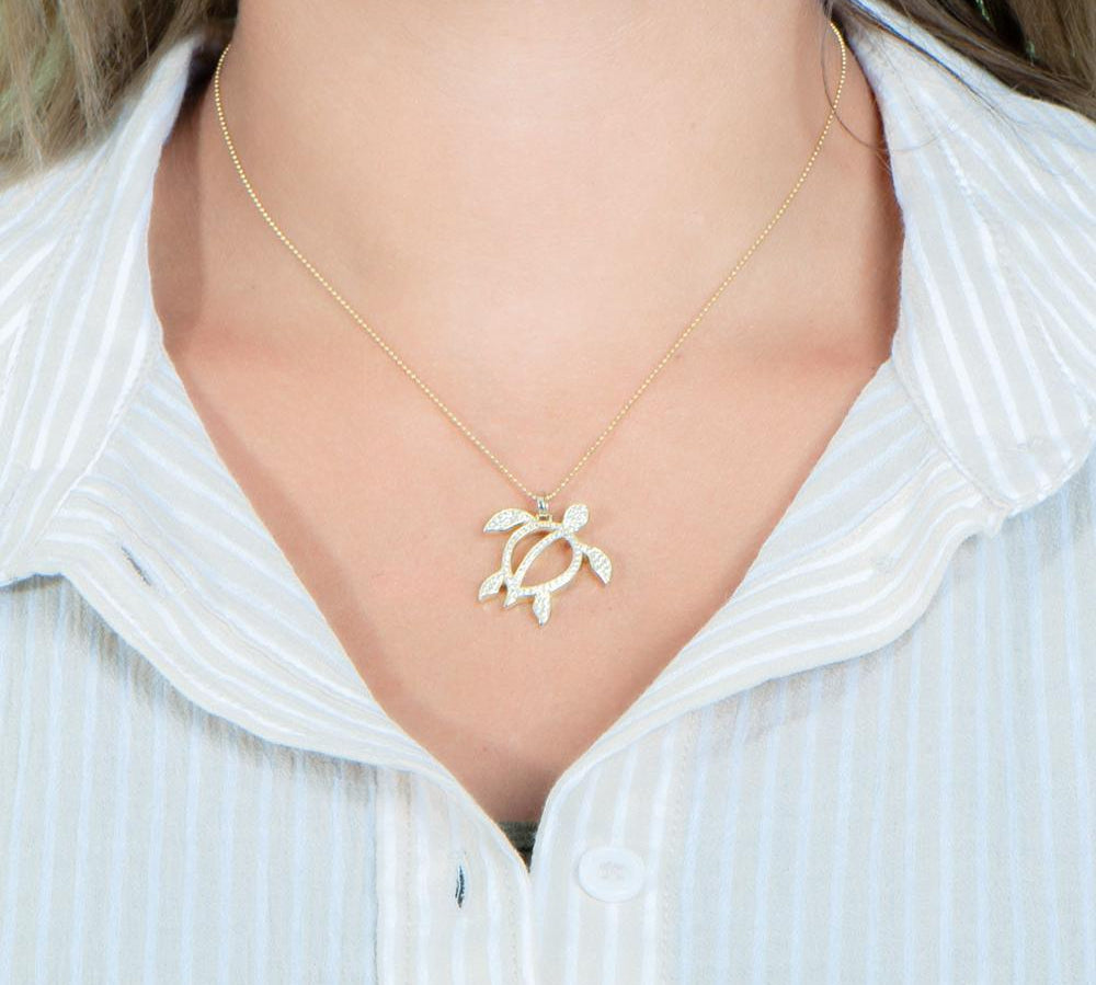 In this photo there is a model with blonde hair and a tan and white striped shirt, wearing a yellow gold sea turtle pendant with topaz gemstones.