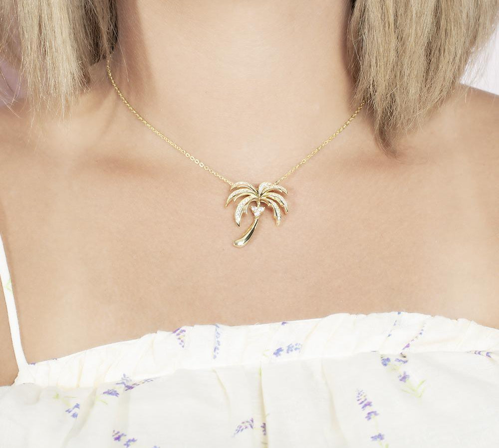 In this photo there is a model with blonde hair and a white shirt with flowers, wearing a yellow gold palm tree pendant with topaz gemstones.