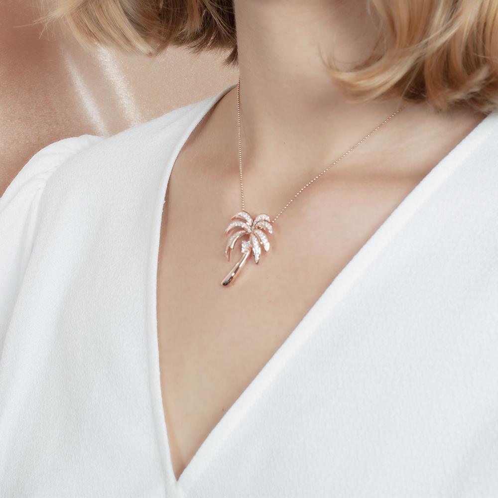In this photo there is a model turned slightly to the left with blonde hair and a white shirt, wearing a rose gold palm tree pendant with topaz gemstones.