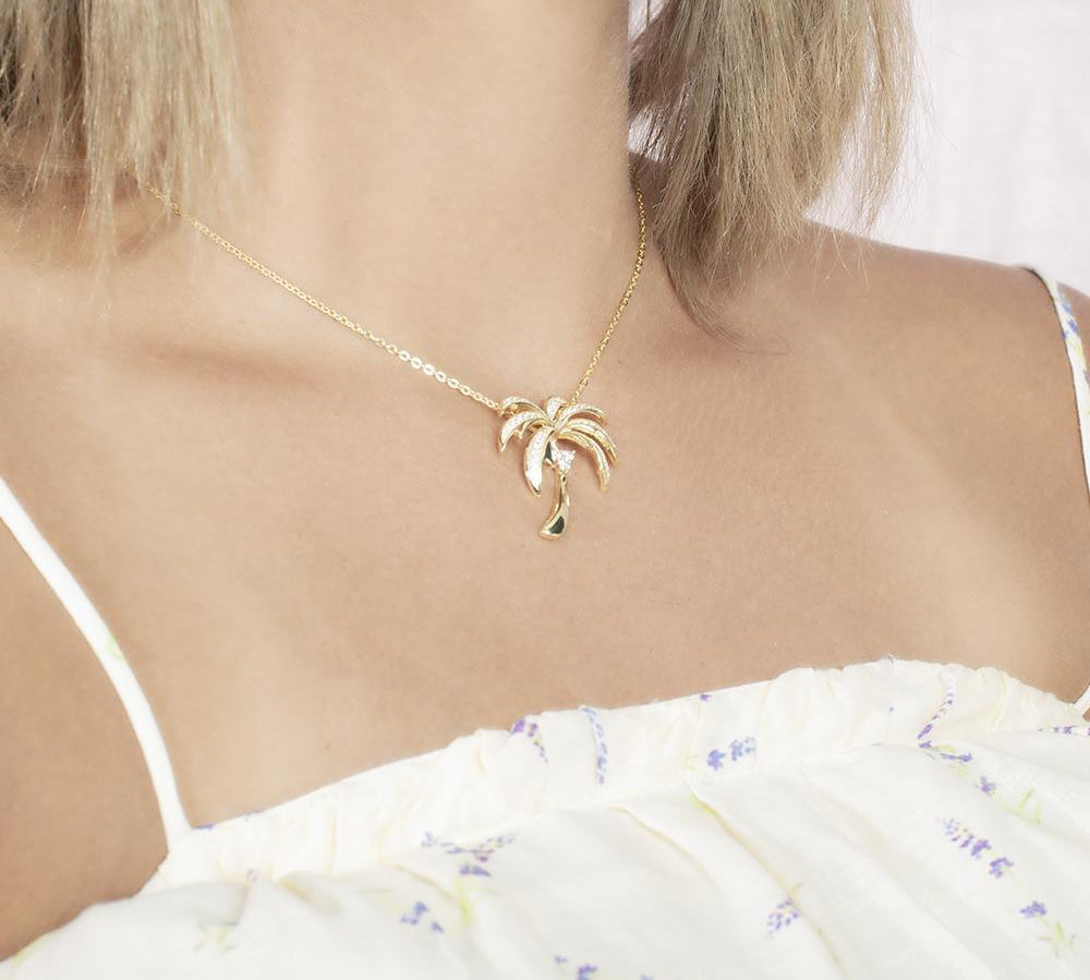In this photo there is a model turned slightly to the right with blonde hair and a white shirt with flowers, wearing a yellow gold palm tree pendant with topaz gemstones.