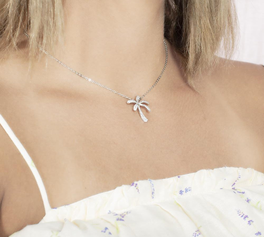 In this photo there is a model turned slightly to the right with blonde hair and a white shirt with flowers, wearing a white gold palm tree pendant with topaz gemstones.