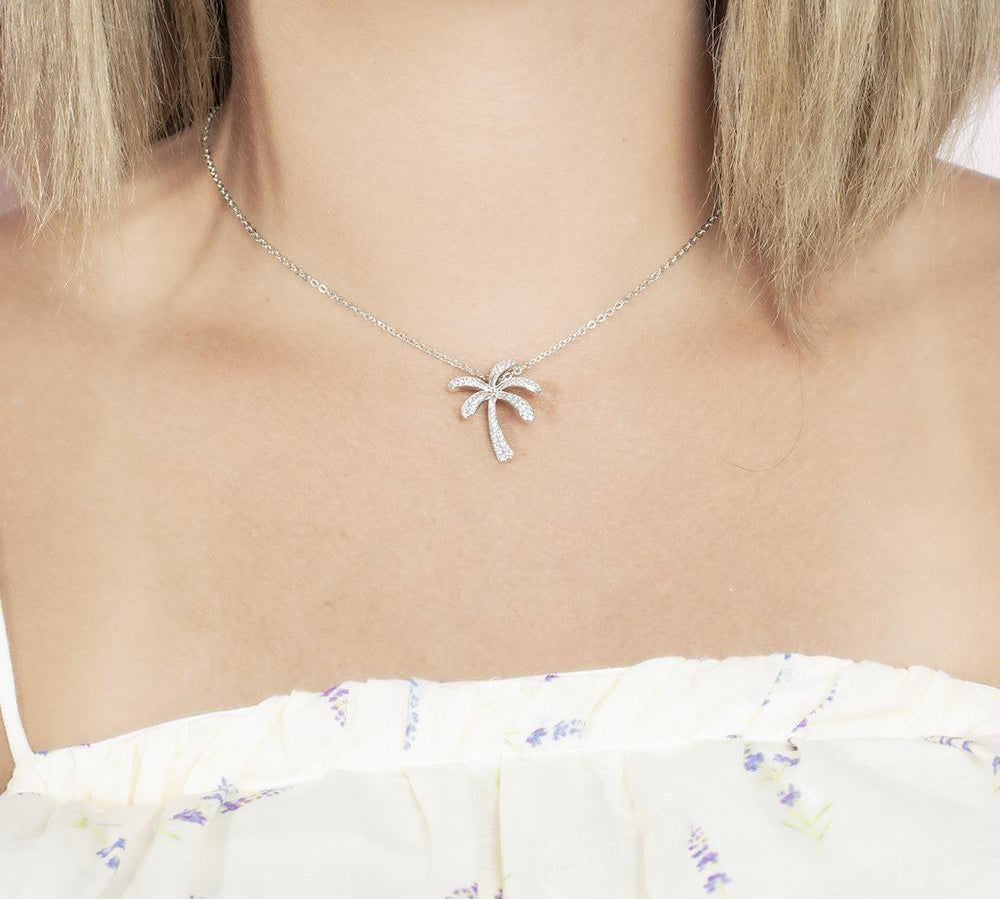 In this photo there is a model with blonde hair and a white shirt with flowers, wearing a white gold palm tree pendant with topaz gemstones.