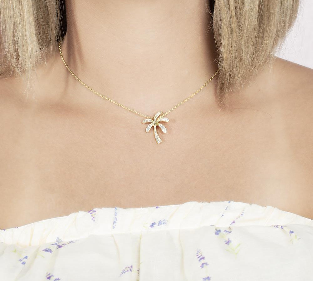 In this photo there is a model with blonde hair and a white shirt with flowers, wearing a yellow gold palm tree pendant with topaz gemstones.