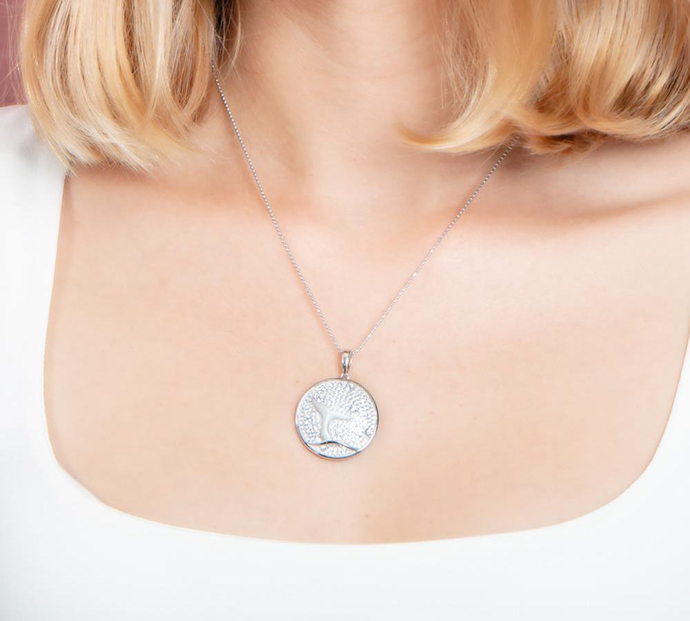 The picture shows a model wearing a 925 sterling silver, white gold vermeil, pavé whale tail medallion pendant with topaz.
