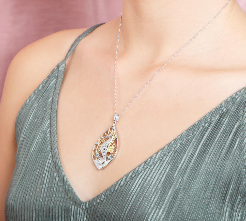 The picture shows a model wearing a two-tone white and yellow gold sea horse mandorla pendant with aquamarine and topaz.