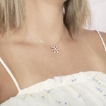 In this photo there is a model with blonde hair and a white shirt with purple flowers turned to the right, wearing a rose gold tiare gardenia pendant with topaz gemstones.